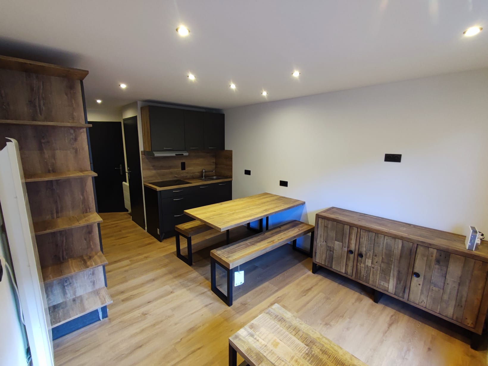 Appartement à Méribel moderne avec meubles noirs et bois, table rectangulaire, bancs et étagères murales, éclairage encastré.
