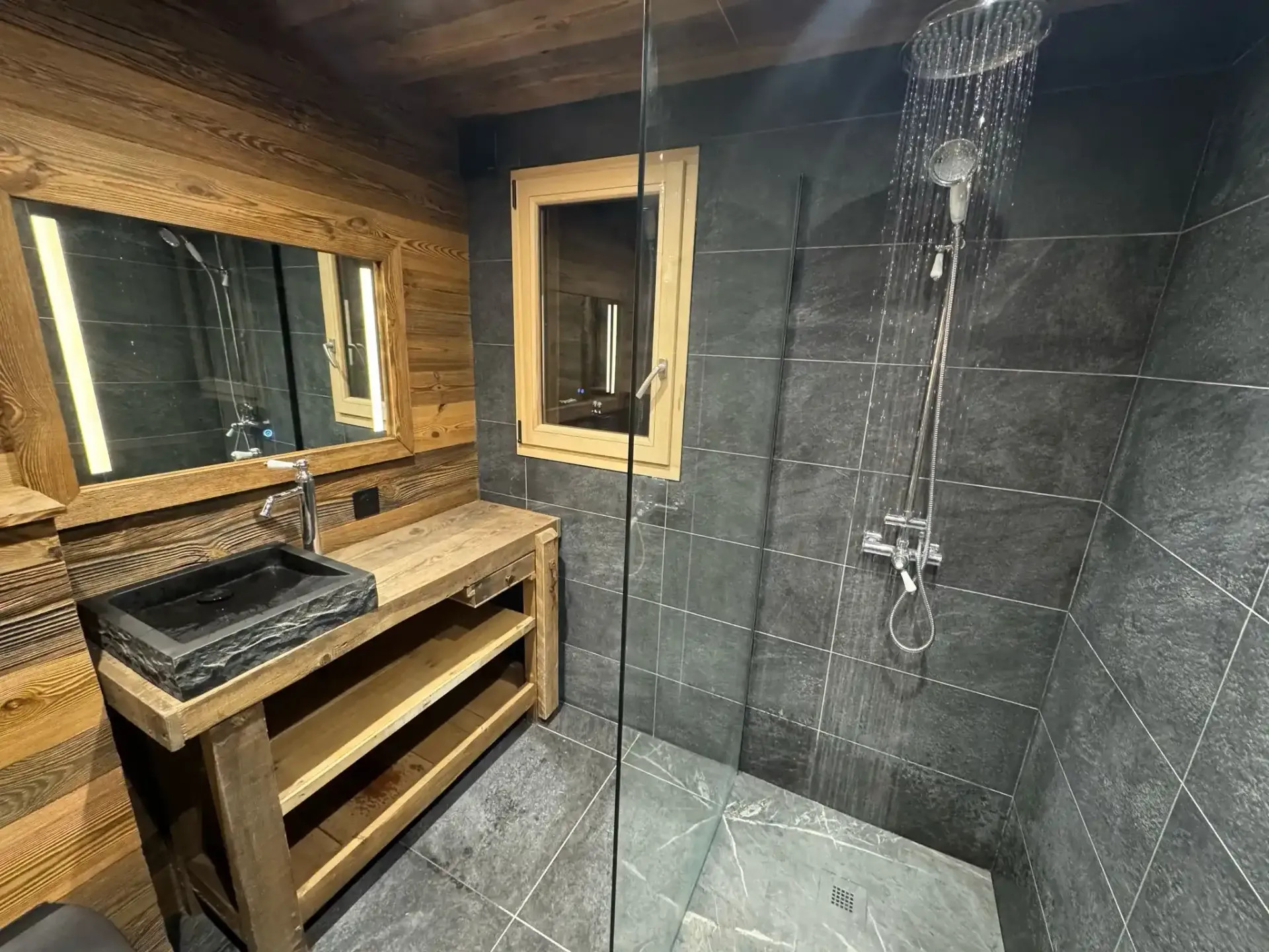 Salle de bain moderne avec douche vitrée, robinetterie chromée, carrelage gris et meuble en bois avec vasque noire.